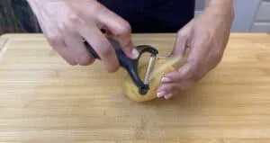 Person peeling a potato with a peeler on a wooden cutting board, showcasing food preparation techniques.
